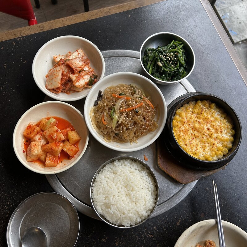 An overhead spread of colorful Korean banchan side dishes -- kimchi, pickled radish, japchae, corn cheese, and steamed rice