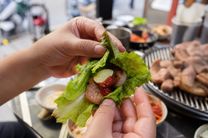 Hands folding a fresh lettuce ssam wrap filled with grilled pork, garlic, and ssamjang sauce