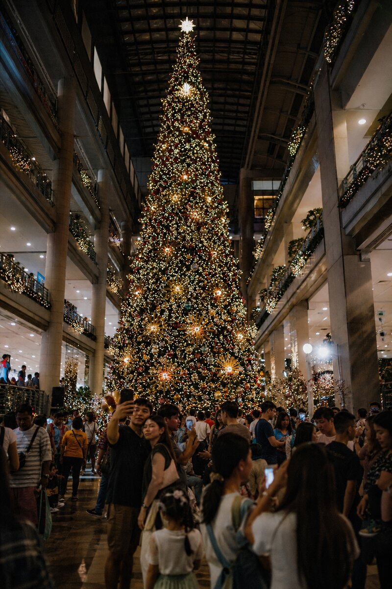 A towering Christmas tree glowing with thousands of lights inside a Metro Manila mall atrium at night, crowds taking photos