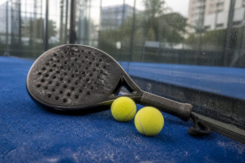 A padel racket and yellow balls resting on the blue court surface near the glass wall